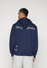 Marineblaue Kapuzenjacke mit Kordelzug und gesticktem "New York Yankees" auf dem Rücken. Gerippte Bündchen und Saum, ausgestattet mit einem Logoschild an der Seite.