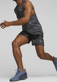 Pantaloni sportivi neri con un motivo testurizzato e canotta grigia, con logo, abbinati a scarpe da allenamento blu. Fisico muscoloso in movimento.