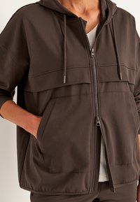Brun zip-up hoodie med hætte, to forlommer og en glat tekstur. Har et splittet design med lysere sektioner ved skuldrene.