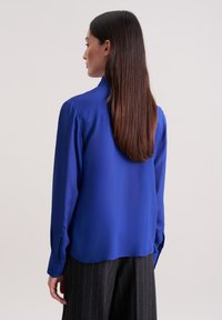 Blaue Seidenbluse mit glatter Textur, langen Ärmeln und Knopfmanschetten, kombiniert mit schwarzen Nadelstreifen-Hosen, von hinten gezeigt.