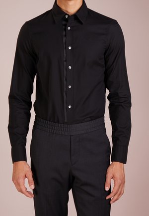 Mann trägt ein eng geschnittenes schwarzes Hemd mit Knöpfen und Kragen sowie dunkle, hoch taillierte Hosen mit elastischem Bund.