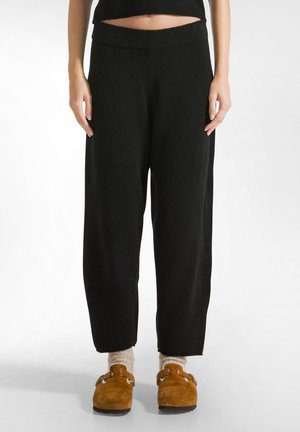Zwarte, wijde broek met een zachte textuur en een elastische tailleband, gecombineerd met bruine suède klompen en lichte sokken.