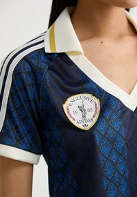 Adidas-tröja med ett svart och blått bladmönster, vit och gul krage med randiga detaljer, och en framträdande broderad logotyp.