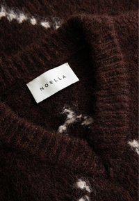 Smeđi pleteni pulover s bijelim uzorcima, sa V-izrezom i rebrastom teksturom. Na etiketi piše "NOELLA" crnim serifnim fontom.