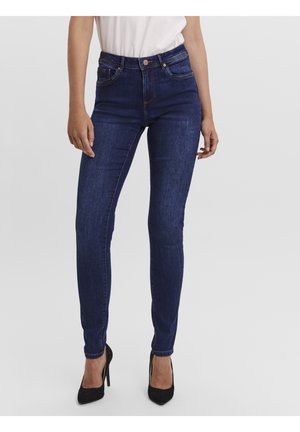 Femme portant un jean skinny bleu foncé, un haut blanc et des talons hauts noirs, debout devant un fond uni clair.