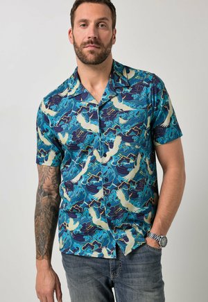 Uomo con camicia blu a maniche corte con motivi bianchi di uccelli e onde, braccio sinistro tatuato, jeans blu e orologio da polso argento.