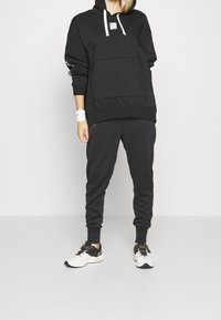 Musta huppari, jossa on etutasku ja valkoiset kiristysnyörit, yhdistettynä mustiin verkkareihin ja valkoisiin urheilukenkäsillä, joissa on mustia yksityiskohtia.