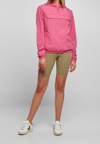 Kobieta w różowej kurtce z długim rękawem, beżowo-różowych spodenkach rowerowych i białych trampkach, stojąca na tle jednolitego, jasnoszarego tła.