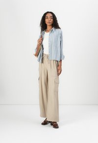 Blauw-wit gestreept overhemd met knopen, gecombineerd met beige wijde cargobroeken. Accessoires omvatten een bruine tas en donkerbruine schoenen.