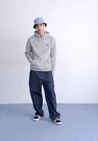 Felpa grigia con cappuccio e cordino, cappello a secchio blu, jeans blu larghi e sneaker nere con dettagli gialli, su uno sfondo neutro.