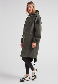 Abrigo impermeable verde oliva con capucha, bolsillo con cremallera en el frente, puños elásticos y dobladillo ajustable, combinado con leggings negros y zapatillas.