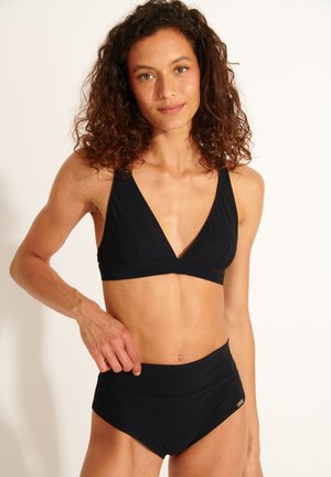 Frau mit lockigem Haar, die einen schwarzen zweiteiligen Badeanzug mit V-Ausschnitt-Oberteil und hoch taillierten Hosen trägt, stehend vor einem weißen Hintergrund.