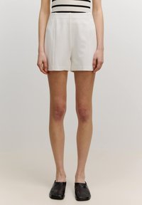Weiße Baumwollshorts mit hoher Taille, versehen mit Falten und einem geraden Schnitt. Kombiniert mit einem schwarz-weißen gestreiften Oberteil und schwarzen flachen Schuhen.
