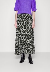 Une personne portant un pull violet, une jupe midi florale noire et des bottines en cuir noires, se tenant contre un fond clair plain.