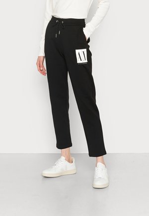 Persona que lleva pantalones negros con cordón y logo blanco de Armani Exchange en el muslo y zapatillas blancas, de pie con una mano en el bolsillo.