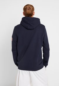 Mörkblå huvtröja med ribbade manschetter och nederkant, med en liten amerikansk flagga på vänster ärm. Mjuk textur.