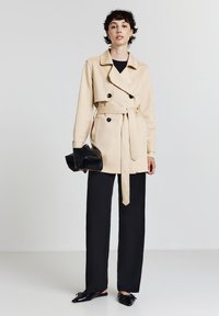 Giovane donna che indossa un trench beige con cintura, pantaloni neri a gamba larga, in mano una clutch nera, in piedi contro uno sfondo bianco uniforme.