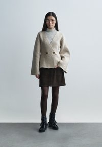 Béžový kabát ze směsi vlny s volánovými rukávy a dvojitými zlatými knoflíky, doplněný proužkovaným rolákem, hnědou sukní a černými šněrovacími botami.