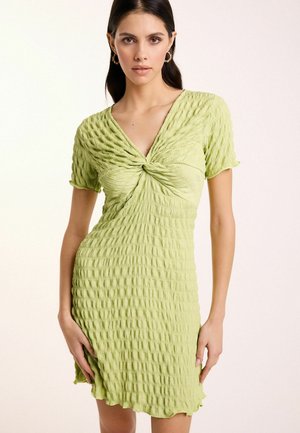 Hellgrünes Kleid aus strukturiertem, zerknittertem Stoff, mit V-Ausschnitt, kurzen Ärmeln und einem verdrehten Vorderteil, das knapp über dem Knie endet.