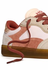 Zapatilla con una parte superior de gamuza y cuero en colores rosa, beige y óxido. Presenta cordones redondos de color óxido y una suela de goma texturizada.