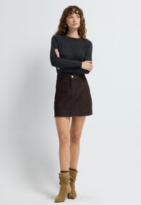 Vrouw met krullend haar in een zwart langemouwen trui, een donkerbruine mini-jurk en slappe tan enkelboots, staande met gekruiste armen.