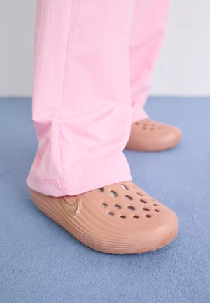 Sabots Nike en plastique beige avec perforations, côtés texturés et un swoosh doré. Portés avec un pantalon rose sur un sol bleu.