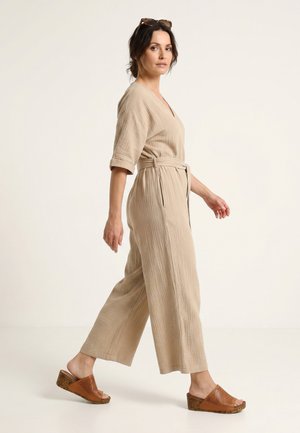 Kvinde går iført en beige jumpsuit med bælte, brune kilehælssandaler og solbriller, der hviler på hendes hoved mod en ensfarvet baggrund.