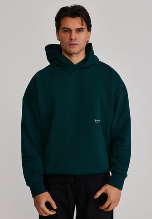 Homme portant un sweat à capuche vert foncé avec poche avant et petit logo blanc, debout devant un fond gris clair uni.