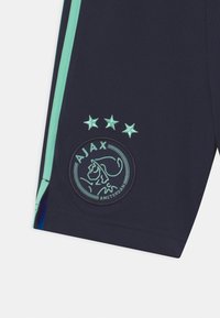 Laivastonsiniset urheilushortsit vaaleanturkooseilla sivuraidoilla. Etupuolella kirjailu Ajax Amsterdamin logo ja kolme tähteä sen yläpuolella. Pehmeä kangas.
