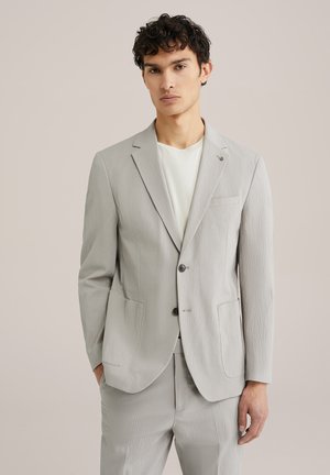 Giacca di suit grigia chiara con texture, dotata di due tasche frontali e chiusura a un bottone, abbinata a una maglietta leggera sotto.