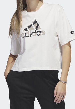 Personne portant un t-shirt court blanc Adidas avec un logo noir et beige, un pantalon noir, un bracelet et des bagues, debout avec une main dans la poche.