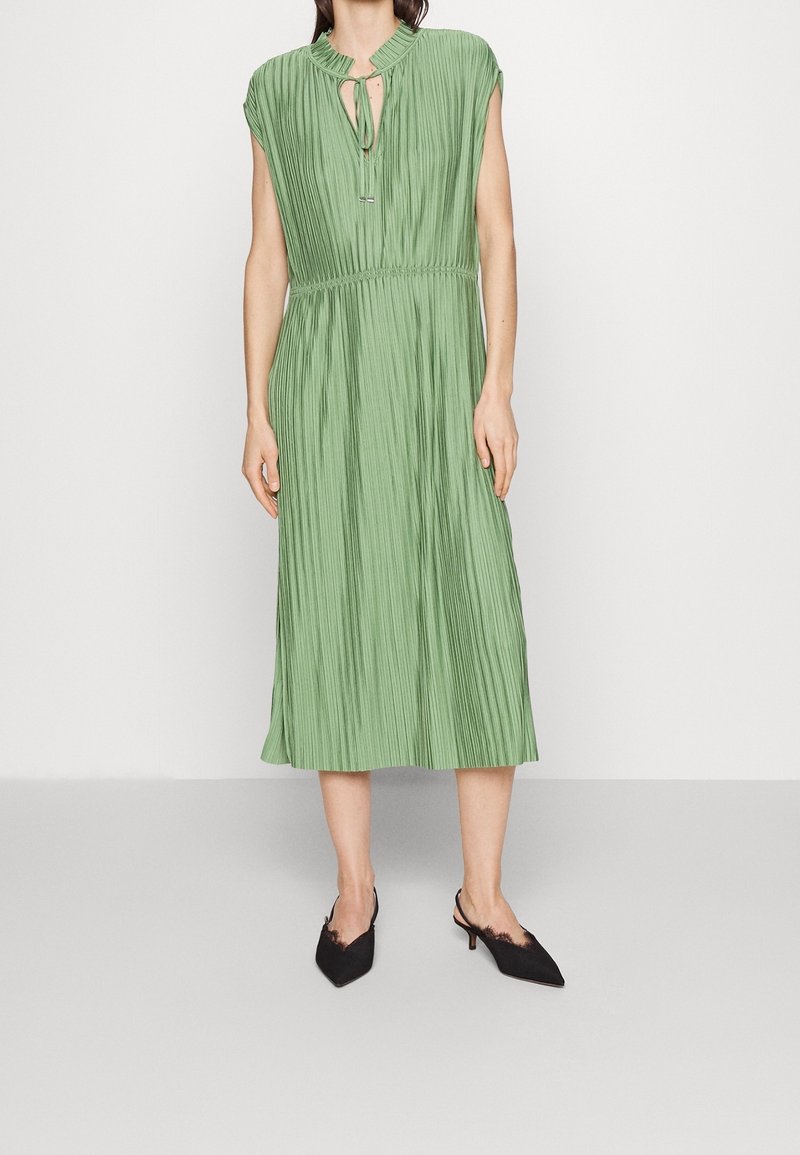 Faltenrockkleid in Grün, Midi-Länge, mit kurzen Ärmeln, Schlüsselloch-Ausschnitt und elastischem Bund. Getragen mit schwarzen Slingback-Absätzen.