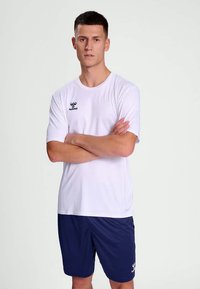 Biele krátke športové tričko s okrúhlym výstrihom, s čiernym logom na ľavej hrudi, v kombinácii s tmavomodrými šortkami.