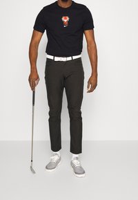 Svart t-shirt med grafisk design, mörka byxor, vit bälte och grå sportskor. Golfklubba hålls i ena handen.