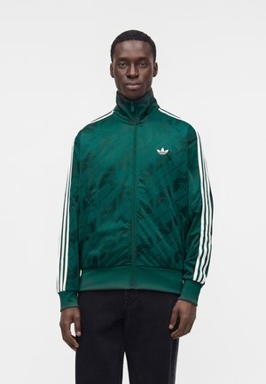 Άνδρας που φοράει πράσινο αθλητικό μπουφάν Adidas με λευκές ρίγες στα μανίκια και λογότυπο στο στήθος, στέκεται μπροστά σε ένα απλό φωτεινό φόντο.