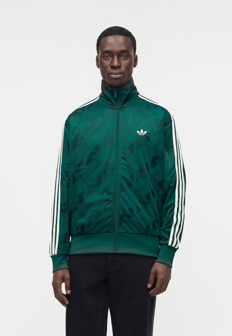 Hombre vistiendo una chaqueta deportiva verde Adidas con rayas blancas en las mangas y logo en el pecho, de pie frente a un fondo claro y liso.