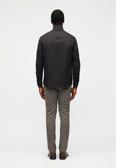 Camisa preta de manga comprida com gola, combinada com calças cinzentas com padrão e sapatos castanhos, vista de costas. Design e ajuste simples.