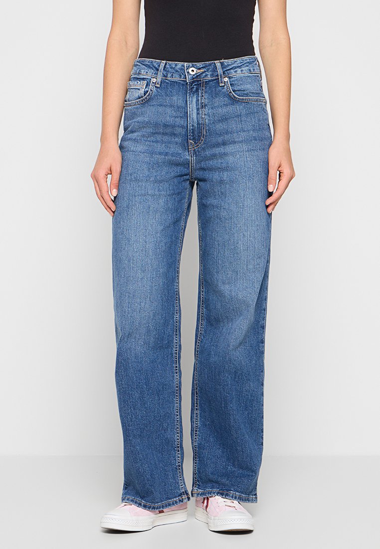 Weite blaue Jeans aus Denim mit hoher Taille, Vorderertaschen und Knopfverschluss. Getragen mit weißen Sneakers.