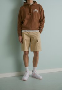 Sudadera con capucha marrón con bolsillo delantero y logo de Champion, combinada con pantalones cortos cargo beige y zapatillas blancas con detalles en rosa.