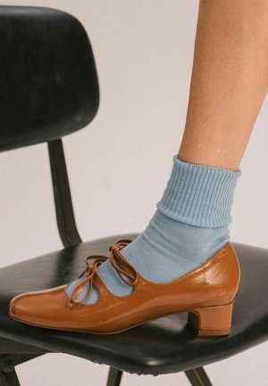 Pied portant une chaussette côtelée bleu clair et une chaussure Mary Jane marron à petit talon repose sur un siège de chaise noir.