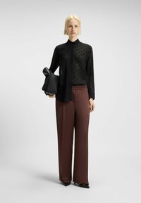 Vrouw met kort blond haar, gekleed in een doorschijnend zwart patroon shirt, bruine wijde broek, zwarte hakken en houdt een grote zwarte clutch vast.