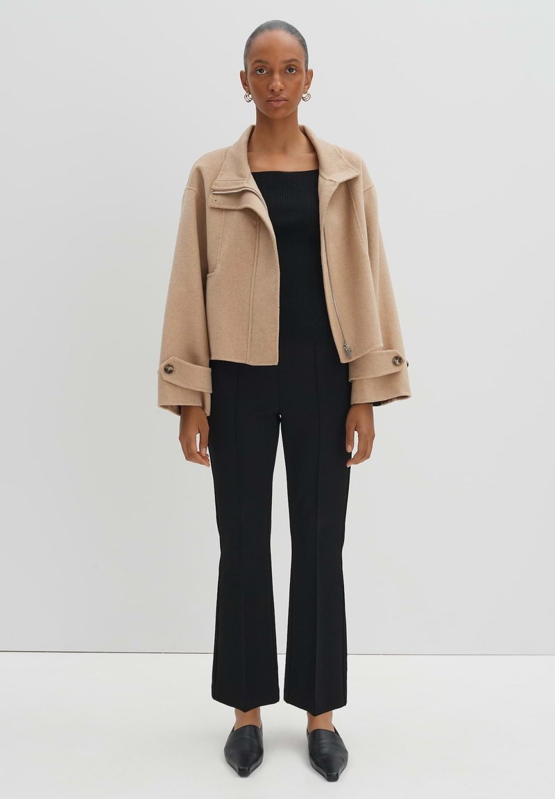 Beige cropped Jacke mit strukturiertem Kragen und Knopfdetails, kombiniert mit einem taillierten schwarzen Rippentop und weit geschnittenen schwarzen Hosen.