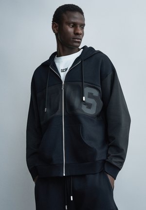 Jeune homme portant un sweat à capuche noir zippé avec de grandes lettres en relief « GCDS », assorti d'un pantalon noir assorti, regardant vers sa gauche.