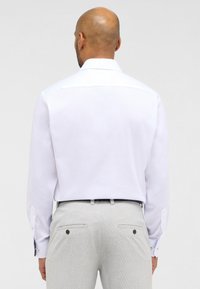 Camicia bianca a maniche lunghe con una texture liscia, caratterizzata da un colletto classico e dettagli con bottoni a contrasto, abbinata a pantaloni grigi chiari a motivo.