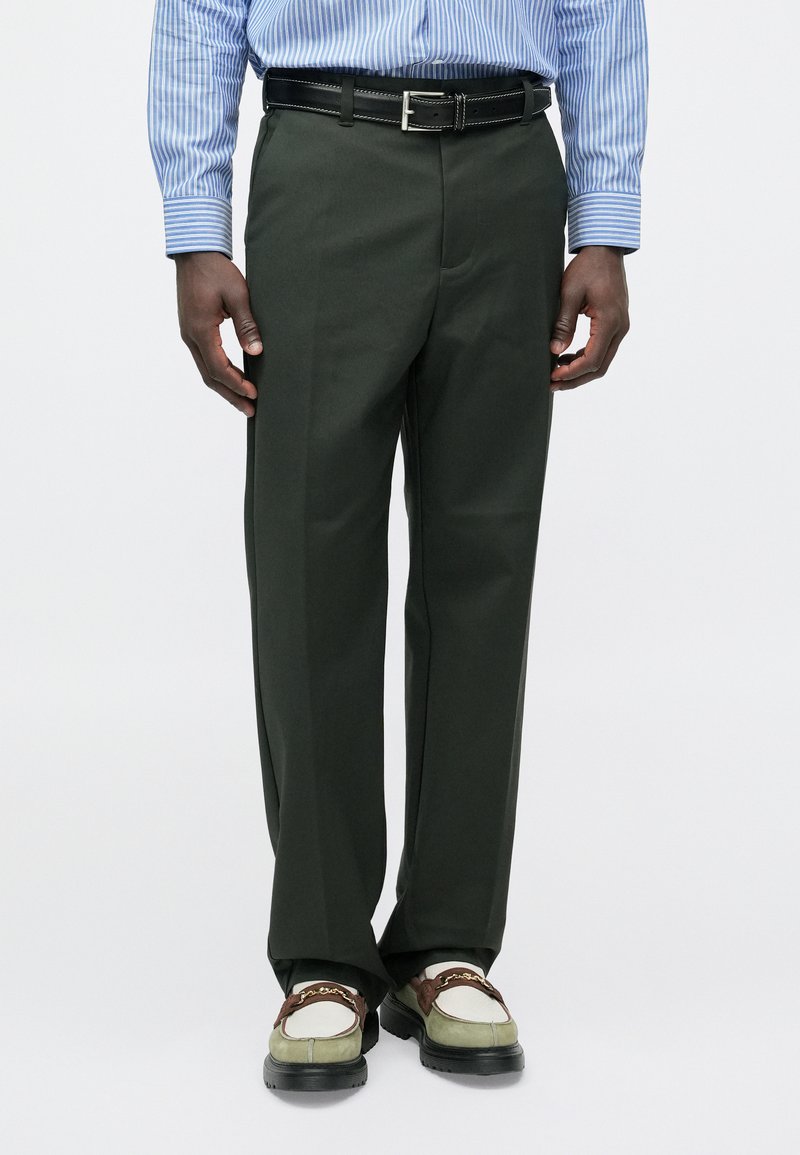 Pantalon vert droit en tissu lisse, avec un pli avant et des poches latérales. Associé à des mocassins marron et verts.