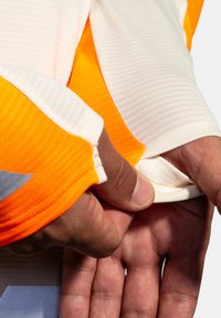 Långärmad plagg med en vit och orange färgsättning, med ribbad textur och åtsittande mudd. En hand håller i tyget vid ärmen.
