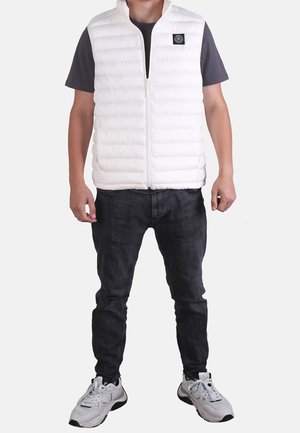 Mężczyzna w białej pikowanej kamizelce na ciemnym krótkim rękawem, czarne dżinsy i białe trampki, stoi na jednolitym tle.