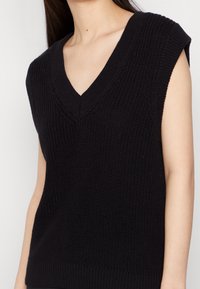 Svart stickad ärmlös topp med V-ringning. Har texturerad tyg och ribbad nederkant. Designad med en avslappnad passform och breda axelöppningar.