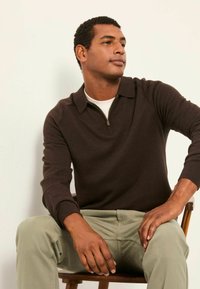 Brauner Strickpullover mit Reißverschluss und Kragen, mit gerippten Bündchen. Getragen über einem weißen Hemd, kombiniert mit hellgrünen Hosen. Sitzt auf einem Hol Stuhl.