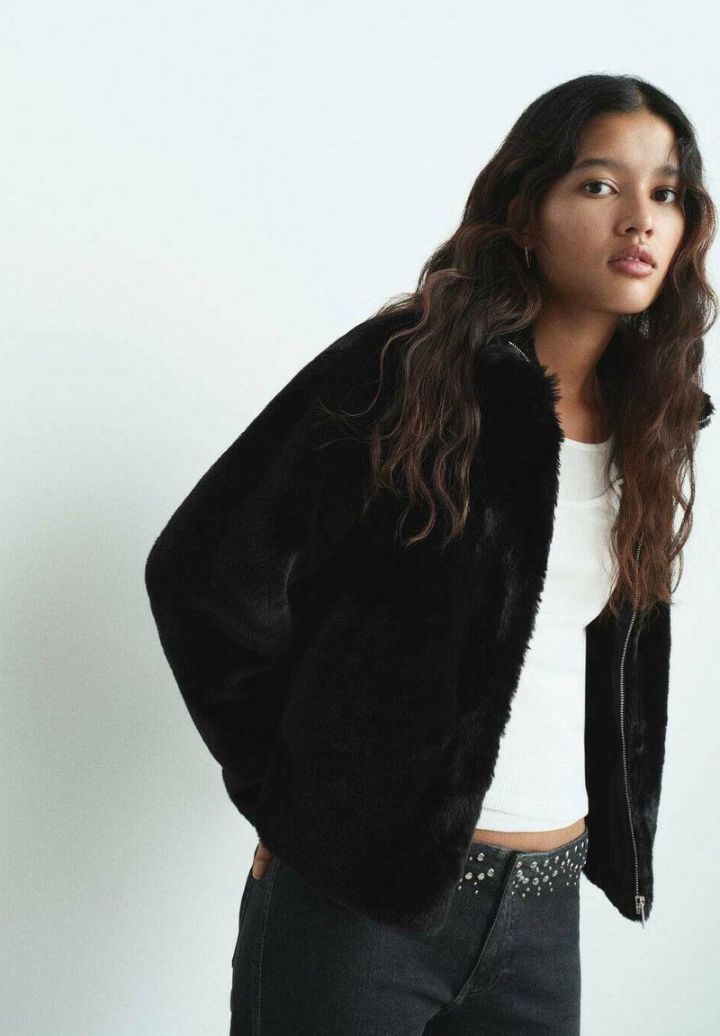 Chaqueta de pelaje sintético negro con cuello alto y cierre de cremallera, combinada con una blusa blanca ajustada y jeans oscuros con acentos de tachuelas.